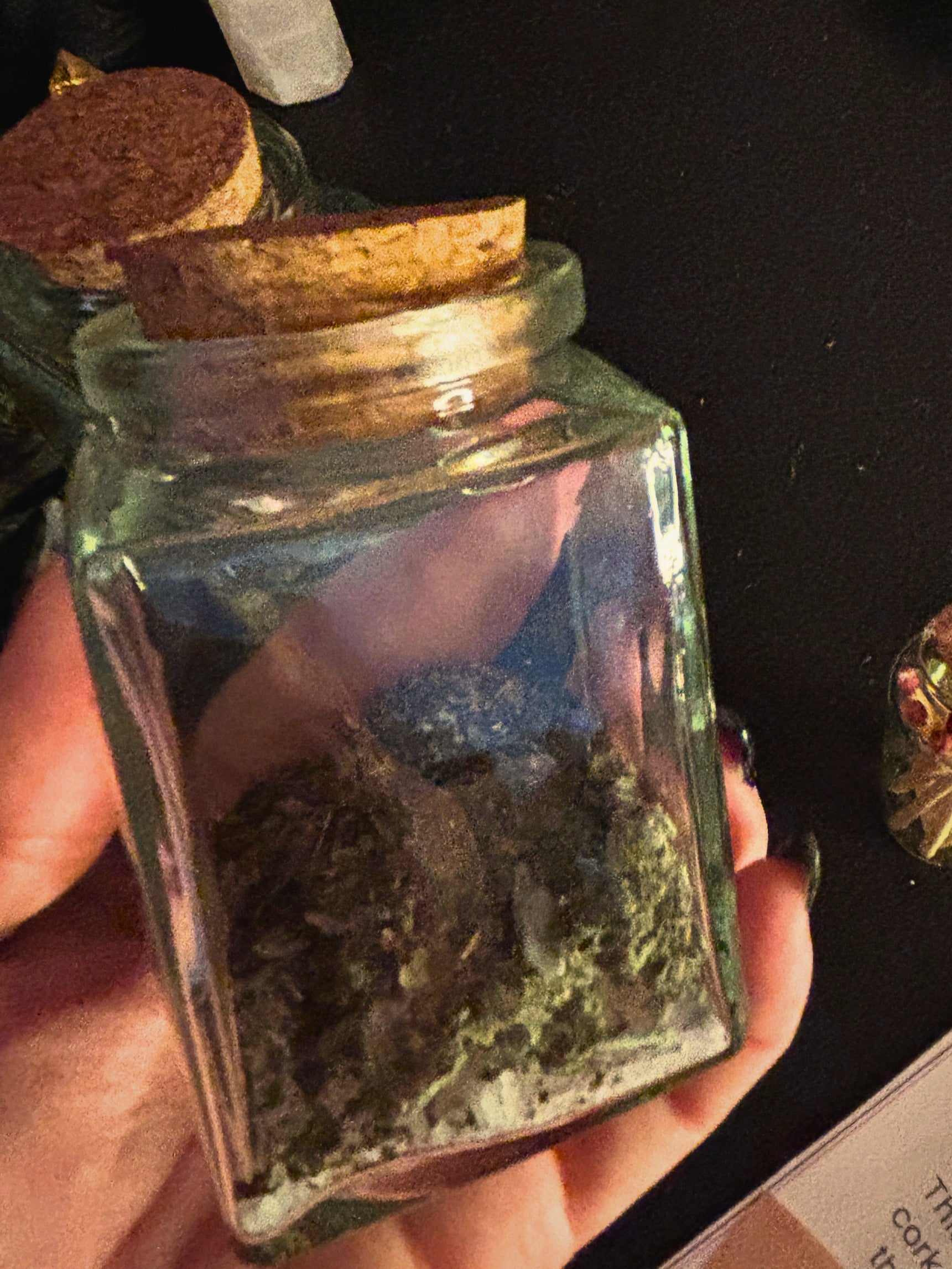 A hand holding a small clear glass jar with a cork stopper, filled with dried green and brown herbal or botanical material, against a dark background.