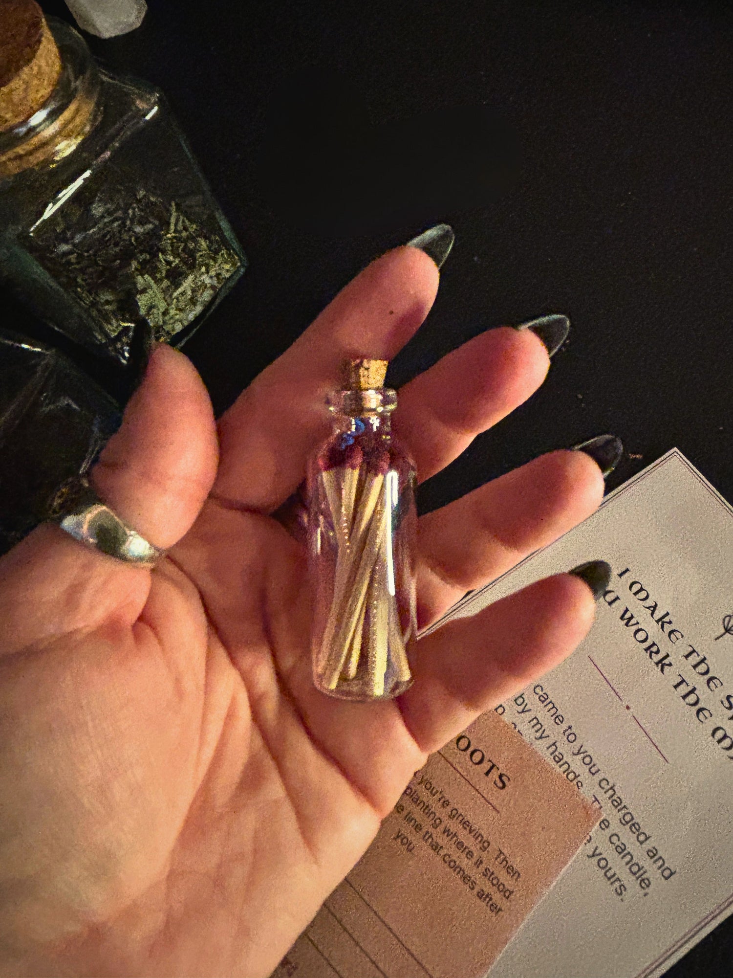 A hand with long dark manicured nails holds a tiny clear glass vial with a cork stopper filled with small wooden matches. The background is dark with a bottle of dried herbs and a printed card visible.