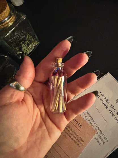 A hand with long dark manicured nails holds a tiny clear glass vial with a cork stopper filled with small wooden matches. The background is dark with a bottle of dried herbs and a printed card visible.