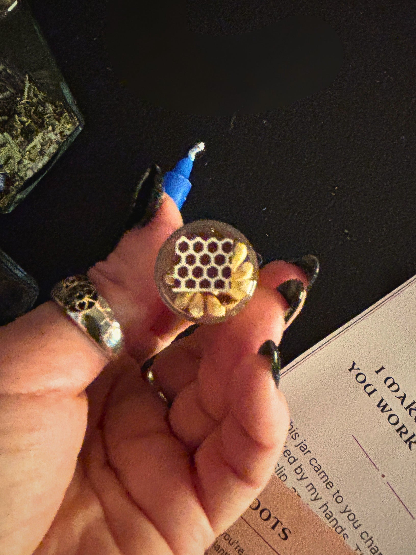 A hand with black nail polish holds a small clear jar containing yellow flower petals and a honeycomb-patterned insert, with a blue applicator tool visible. A dark surface and part of a printed card are in the background.
